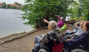 Bewohner*innen der ProCurand Seniorenresidenz Am Straussee stehen am Ufer, zeigen begeistert auf den Straussee und genießen den herrlichen Blick auf die Strausberger Altstadt.