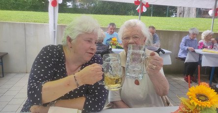 Zwei Seniorinnen stoßen im Garten des ProCurand Seniorenzentrums Am Herzogschloss mit Biergläsern an und genießen den lauen Sommerabend.
