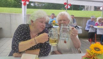 Zwei Seniorinnen stoßen im Garten des ProCurand Seniorenzentrums Am Herzogschloss mit Biergläsern an und genießen den lauen Sommerabend.
