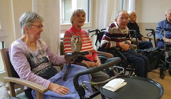 Eine Bewohnerin der ProCurand Seniorenresidenz Bölschestraße hält eine Eule vorsichtig auf dem Arm, während andere Senior*innen die besondere Begegnung interessiert beobachten.