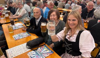 Die Bewohner*innen und eine Mitarbeiterin des ProCurand Pflegestifts Pfarrer Lukas sitzen am Tisch zusammen, lachen und stoßen gemeinsam mit Bierkrügen auf einen fröhlichen Nachmittag beim Chamer Volksfest an.
