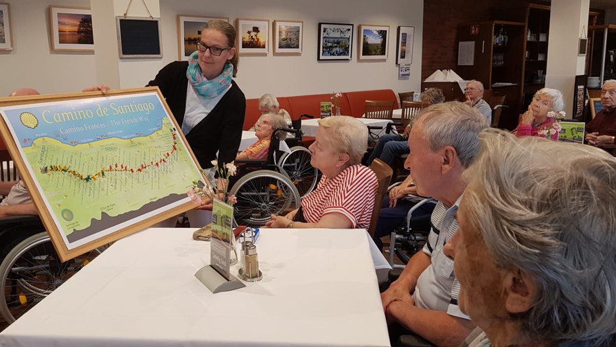 Bewohner*innen der ProCurand Seniorenresidenz Bölschestraße verfolgen aufmerksam eine Lesung, während eine Karte des Jakobswegs gezeigt wird.