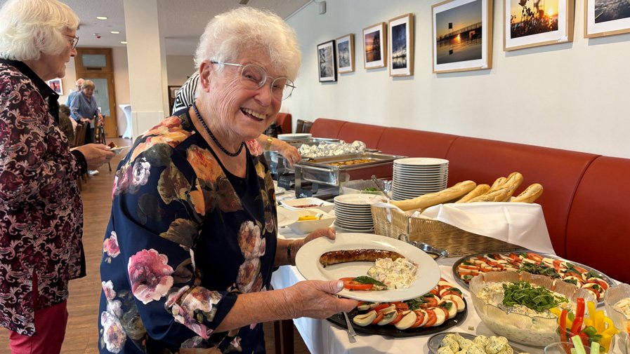 Strahlende Freude am Grillnachmittag – eine Mieterin der ProCurand Seniorenresidenz Bölschestraße lächelt neben dem reich gedeckten Buffet.