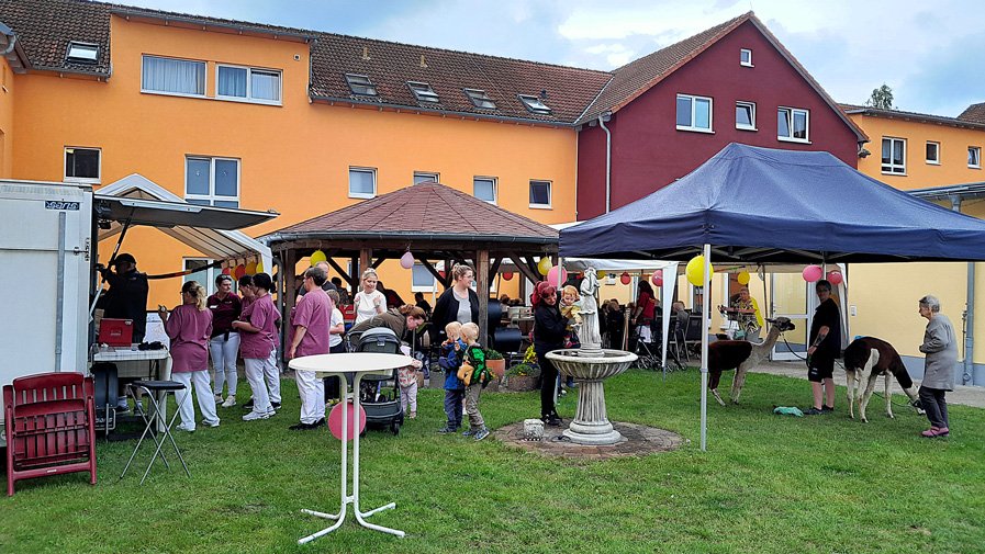Bunte Sommerstimmung im Garten – Bewohnerinnen und Mitarbeiterinnen genießen gemeinsam das Sommerfest. Dekorationen schmücken die Tische, und neugierige Alpakas streifen friedlich durch die Runde, sorgen für Lachen und Gespräche.
