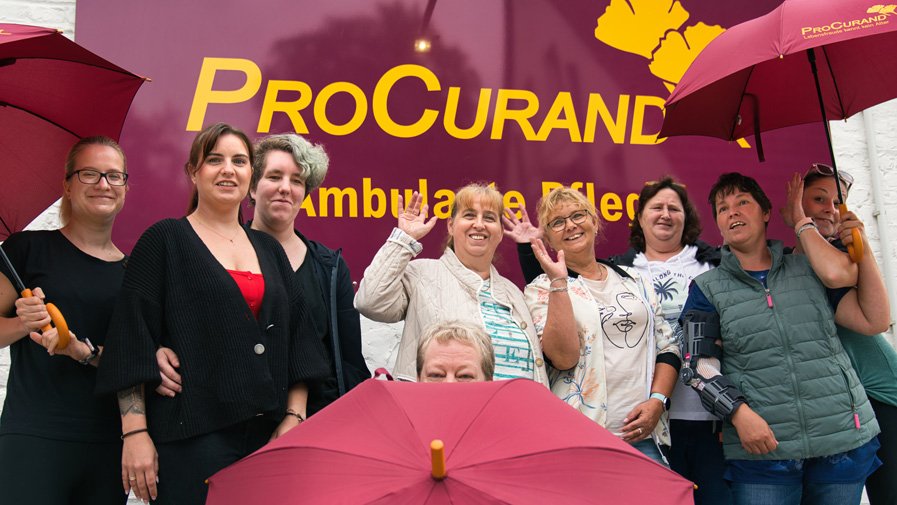 Das Team der ProCurand Ambulanten Pflege Ludwigsfelde steht mit Regenschirmen vor der Einrichtung – gut gelaunt und bereit für einen gemeinsamen Ausflug zum Sound City Festival.