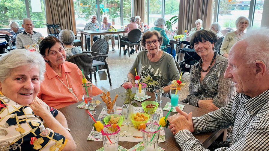 Bewohner*innen und Mieter*innen aus dem Service-Wohnen der ProCurand Seniorenresidenz Havelpalais sitzen in der festlich mit bunten Girlanden geschmückten Cafeteria im Kreis, stoßen fröhlich mit ihren Cocktails an und genießen den gemütlichen Nachmittag.