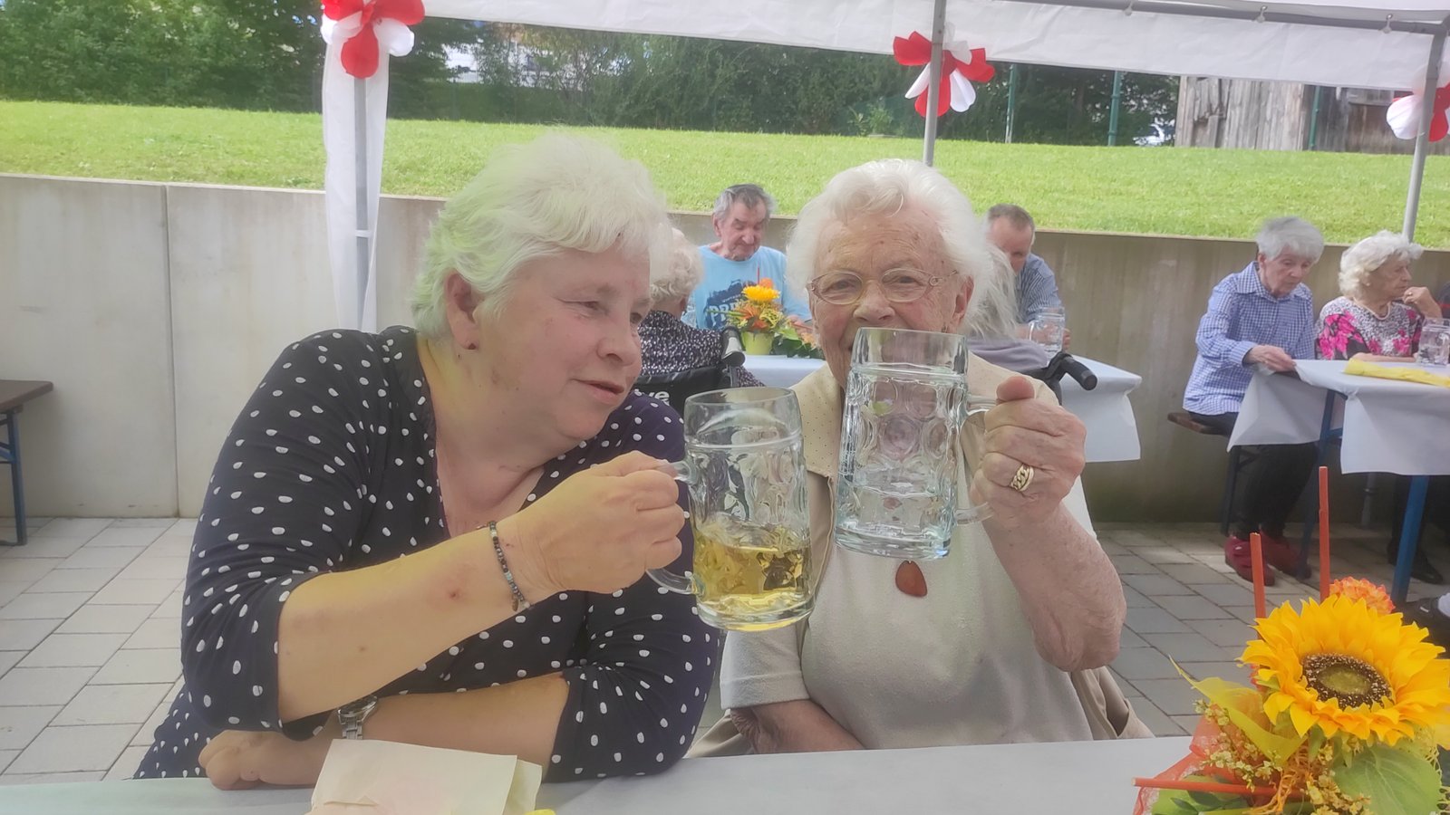 Zwei Seniorinnen stoßen im Garten des ProCurand Seniorenzentrums Am Herzogschloss mit Biergläsern an und genießen den lauen Sommerabend.