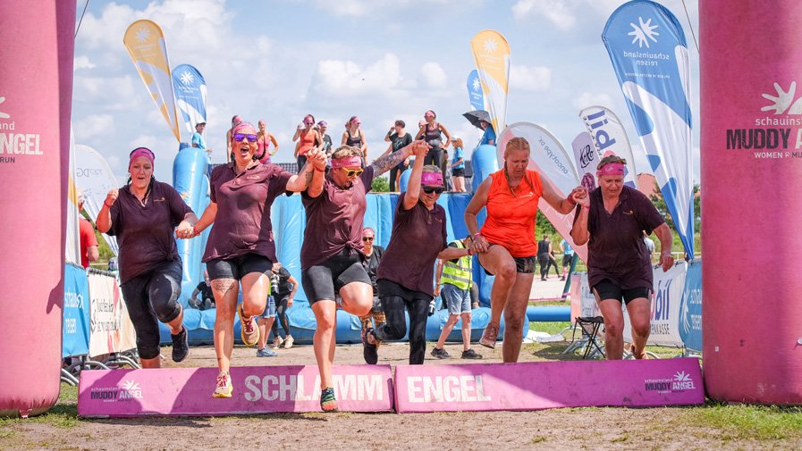 Die Power-Ladies aus der stationären Pflege der ProCurand Seniorenresidenz Am Straussee laufen Hand in Hand lachend über die Ziellinie – voller Schlamm, aber sichtlich stolz.
