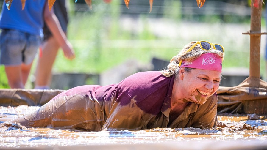 Mit einem breiten Lächeln robbt eine Teilnehmerin durch den Schlammparcours – verschmiert, glücklich und voller Energie.