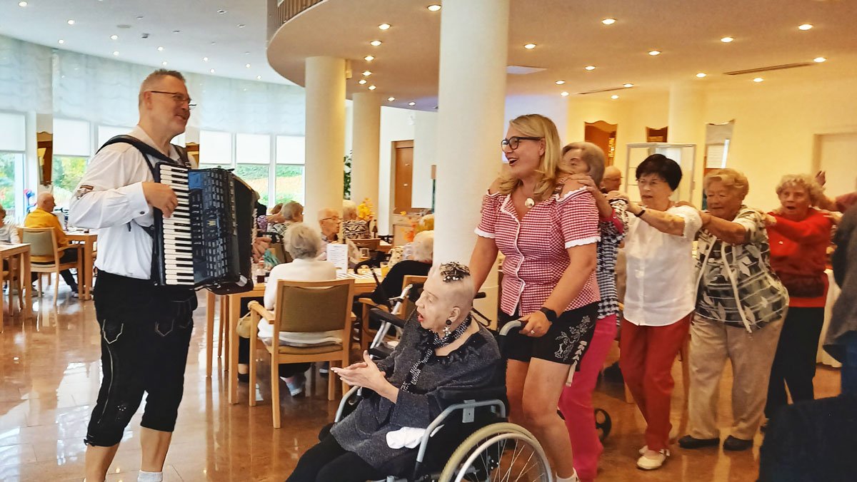 Bewohner*innen der ProCurand Seniorenresidenz Am Straussee tanzen und bewegen sich fröhlich zur Akkordeonmusik von Milan und erleben ein ausgelassenes Oktoberfest.
